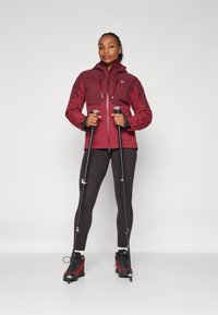 Femme en veste de randonnée rouge et bottes tenant deux bâtons de trekking noirs, se tenant devant un fond uni.