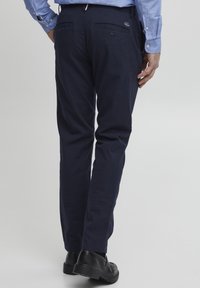 Pantaloni in cotone blu navy con vestibilità dritta, presentano due tasche posteriori con bottoni e un discreto dettaglio del logo. Indossati con scarpe nere.