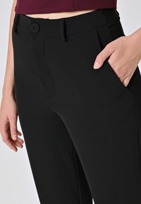 Pantaloni neri con una texture liscia, dotati di una chiusura a un bottone, tasche laterali e un design sartoriale. Vista ravvicinata che mostra i dettagli.