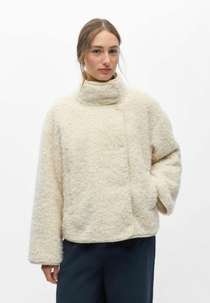 Femme portant une veste polaire crème texturée à col montant avec manches longues et pantalon sombre, debout sur un fond blanc uni.