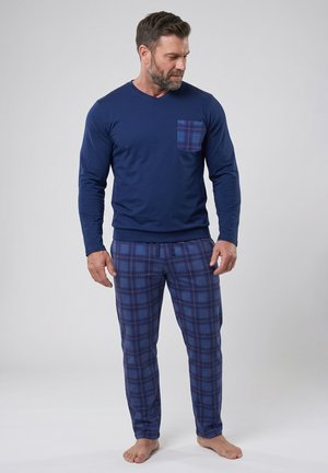 Hombre de pie descalzo vistiendo una camisa azul marino de manga larga con bolsillo de cuadros y pantalones de pijama a cuadros a juego sobre fondo gris.