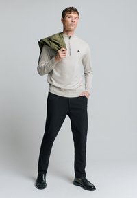 Lichtbeige pullover met ritskraag, gecombineerd met zwarte broeken. Het model houdt een groene jas vast en draagt zwarte instappers. Soepele textuur en aansluitend ontwerp.