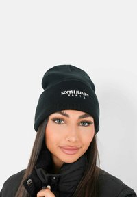 Svart stickad mössa med "SIXTH JUNE" och "PARIS" broderat i vitt. Mjuk textur; figurnära design. Buren med en svart kappa.