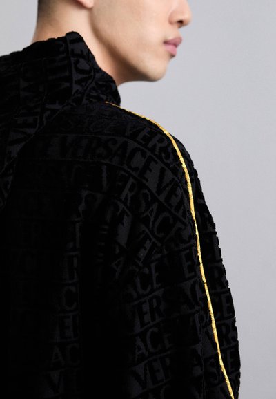 Jachetă din țesătură neagră, prezentând un model de logo Versace reliefat, cu o siluetă slim și un accent auriu pe cusătura umărului.