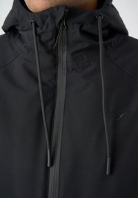 Schwarze, wetterfeste Jacke mit Reißverschluss, verstellbarem Kapuzenzug und subtilen Logo auf der Brust. Glatte Textur.