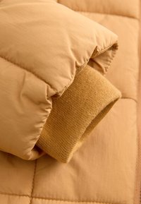 Manche d'une doudoune beige avec un bord-côte en maille à l'intérieur, montrant les coutures et la texture matelassée rembourrée du tissu.