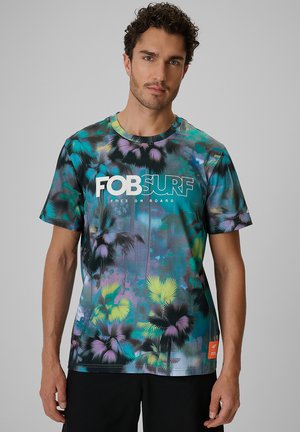 Mężczyzna noszący kolorową koszulkę z krótkim rękawem z tropikalnym nadrukiem palm oraz tekstem "FOB SURF Free On Board" z przodu.