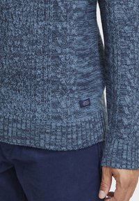 Pulover tricotat de culoare albastru închis cu modele albastre deschise, având o finisare texturată, manșoane cu dantelă și o etichetă mică din material pe tiv.