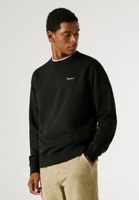 Sweatshirt preto com punhos e bainha canelados, apresentando um pequeno logótipo branco no peito. Combinado com calças de veludo cotelê bege.