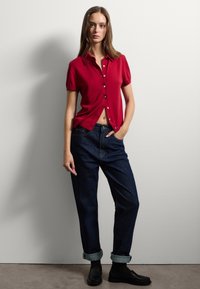Camicia rossa a maniche corte con colletto, abbinata a jeans in denim scuro larghi e mocassini neri, indossata con i polsini arrotolati.