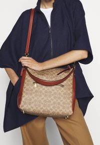 Beige mønstret håndtaske med en buet design, røde læderdetaljer og guldhardware, båret af en person iført en marineblå top.