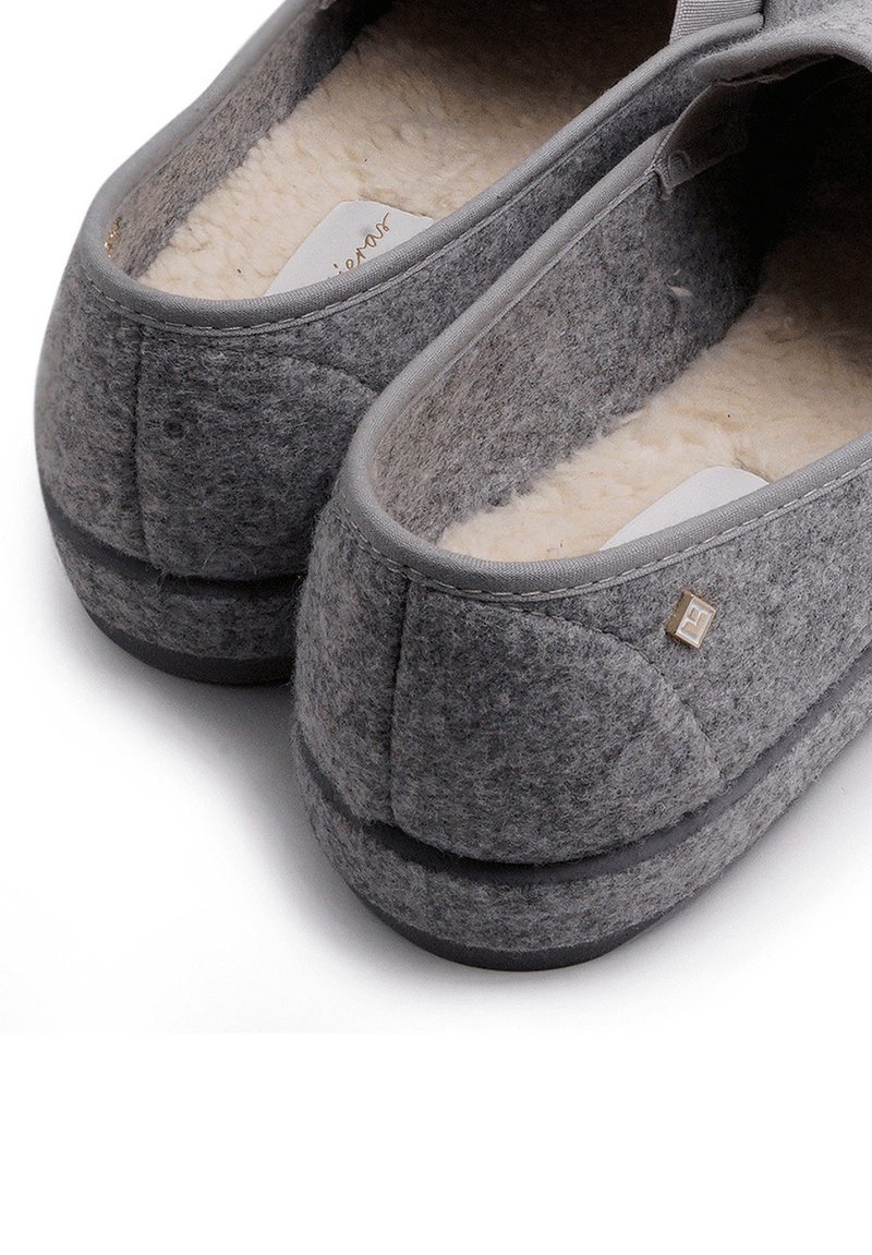 Graue Hausschuhe aus angenehm weichem Material mit einem weichen Innenfutter, runder Form, elastischem Oberteil und einem kleinen Logo-Akzent an der Seite. Verfügt über eine flache Sohle.
