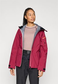 Rød vandtæt jakke med grå accenter, lynlåslommer, justerbare manchetter og hætte. Båret over en lys mønstret sweater og sorte bukser.