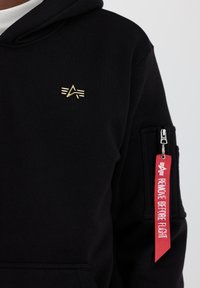 Sort hoodie lavet af bomuld med et guld logo på brystet. Har en lynlåslomme på ærmet med et rødt "REMOVE BEFORE FLIGHT" mærke.