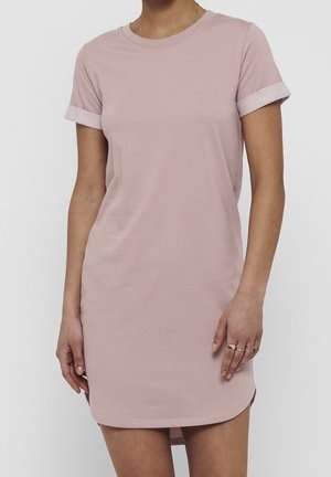 Kobieta ubrana w krótką sukienkę typu T-shirt w kolorze pudrowego różu z podwiniętymi rękawami, nosząca minimalistyczne złote pierścionki na prawej ręce.