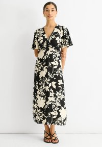 Schwarzes Blumenmaxikleid mit kurzen Ärmeln, das weiße Blumen, einen tiefen V-Ausschnitt und fließenden Stoff zeigt; kombiniert mit schwarzen Sandalen mit Riemchen.