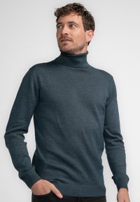 Ciemnoniebieski sweter typu golf w miękkim dzianinowym materiale, z długimi rękawami oraz ściągaczami u mankietów i u dołu. Model stoi z ręką w kieszeni.