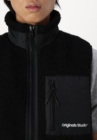 Zwarte fleecevest met hoge kraag, voorzien van een voorrits en een linkerborstzak met rits. Logo in het wit afgedrukt op de zak.