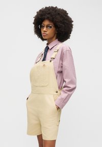 Άτομο με πυκνά, σγουρά μαλλιά φοράει μπεζ κοντό ολόσωμο σορτς, φανελένιο πουκάμισο ανοιχτού μοβ με σχέδια, ναυτικό γραβάτα και γυαλιά με σκουρόχρωμους φακούς, στέκεται με τα χέρια στις τσέπες.