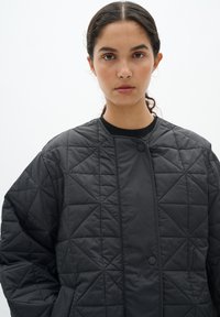 Giacca nera trapuntata con colletto rotondo, chiusura a scatto e texture simile alla seta. Presenta motivi geometrici a diamante su tutta la superficie.