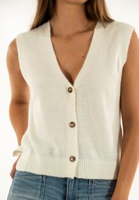 Cardigan bianco senza maniche con scollatura a V profonda, chiusura frontale con bottoni, orlo a coste e una texture morbida e pelosa.