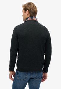 Homme portant un pull gris foncé par-dessus une chemise à carreaux rouge et bleue, associé à un jean bleu, vu de dos sur un fond blanc.
