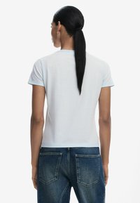 T-shirt ajusté à manches courtes, bleu clair et blanc, avec un design épuré. Le tissu semble doux et est porté avec un jean en denim bleu foncé.