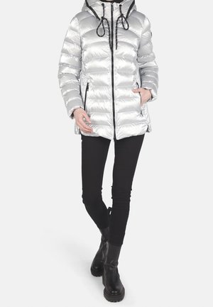 Personne portant une doudoune argentée brillante avec capuche et cordons noirs, pantalon noir et bottines noires épaisses, debout avec une main dans la poche.