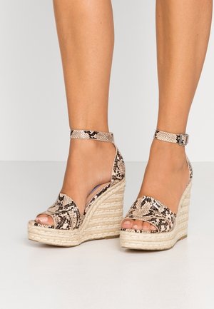 Fødder iført beige og sorte kilehælssandaler med slangeskindsmønster, ankelremme og fletninger på espadrillesåler.