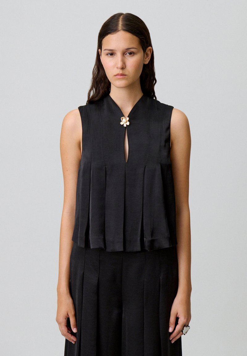 Top negro sin mangas con un diseño plisado, escote en forma de lágrima y un broche floral como acento. Textura suave y tela ligera.