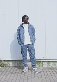 Sporty tracksuit i lys blå med en lynlåsejakke og bukser med elastisk talje. Sammen med grå sneakers og runde solbriller.
