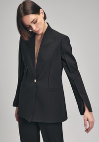 Blazer negro con un profundo escote en V, cierre de un solo botón dorado y elegantes mangas largas. Ajuste entallado con costuras frontales sutiles y bolsillos.