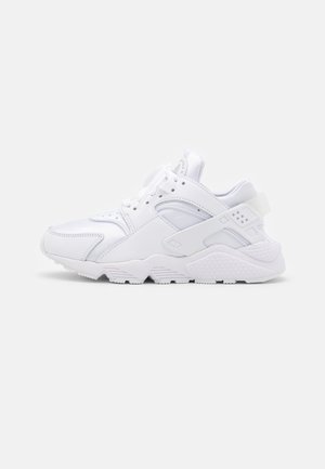Baskets de sport blanches avec panneaux en mesh et cuir, lacets et semelle épaisse sur fond uni gris clair.