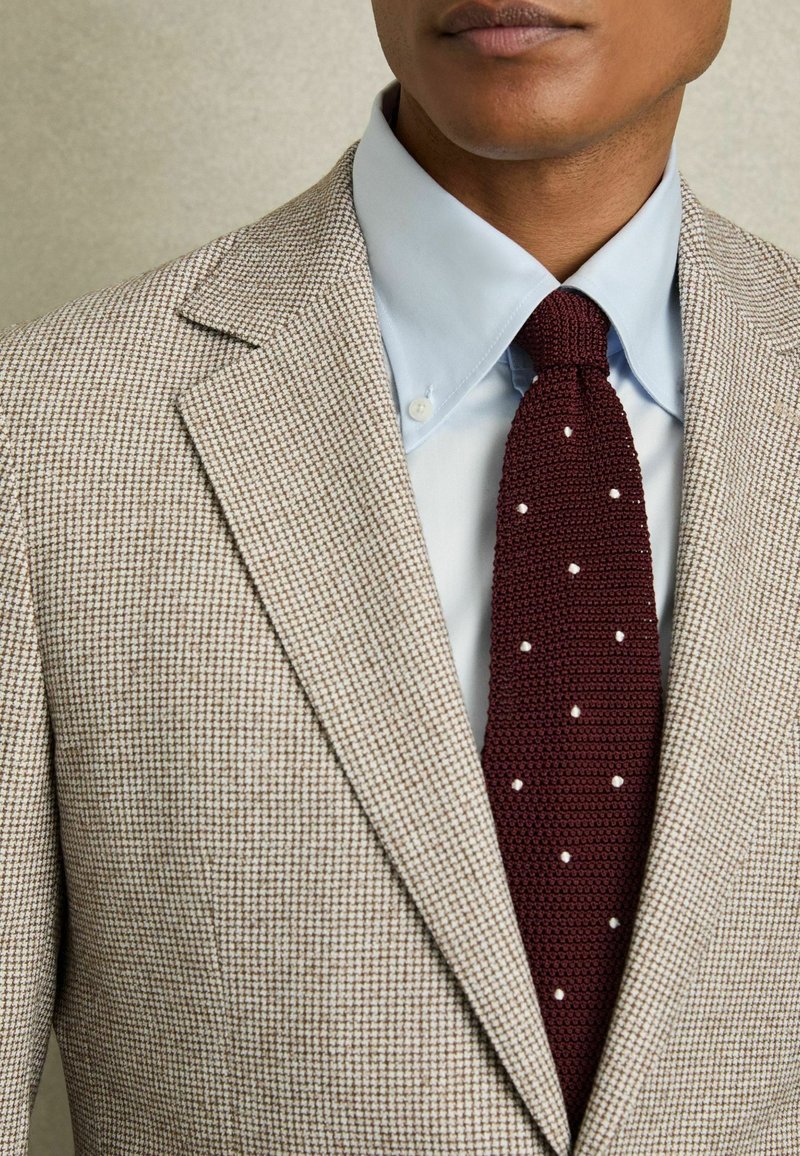 Blazer bej cu carouri, cu rever tip notch, purtat peste o cămașă albastră deschisă. O cravată tricotată în nuanța burgund cu puncte albe adaugă un aspect texturat.