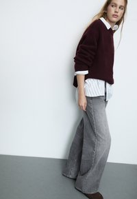 Bordo pleteni pulover preko bijele košulje s prugama, u kombinaciji sa sivim širokim trapericama i smeškim cipelama, na jednostavnoj pozadini.