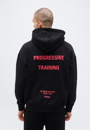 Personne portant un sweat à capuche noir avec le texte rouge "PROGRESSIVE TRAINING" et un texte plus petit "BE YOUR BEST SELF SINCE 1956 BORG" au dos.