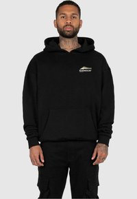 Zwarte hoodie met een voorzak, ribgebreide cuffs en zoom. Bevat een klein wit en gouden logo op de borst. Gemaakt van zacht stof.
