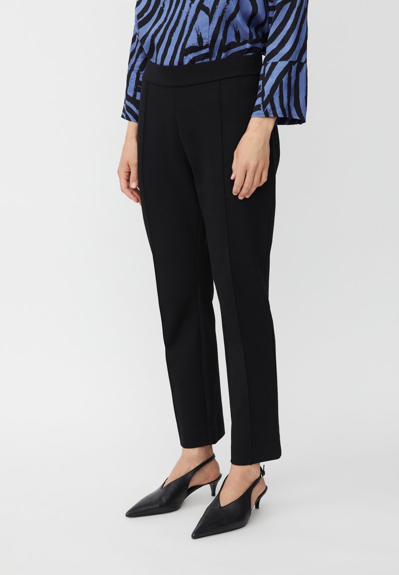 Zwarte op maat gemaakte broek met een zachte textuur, voorzien van een hoge tailleband en een slanke pasvorm die taps toeloopt bij de enkel. G gedragen met zwarte puntige slingbacks.