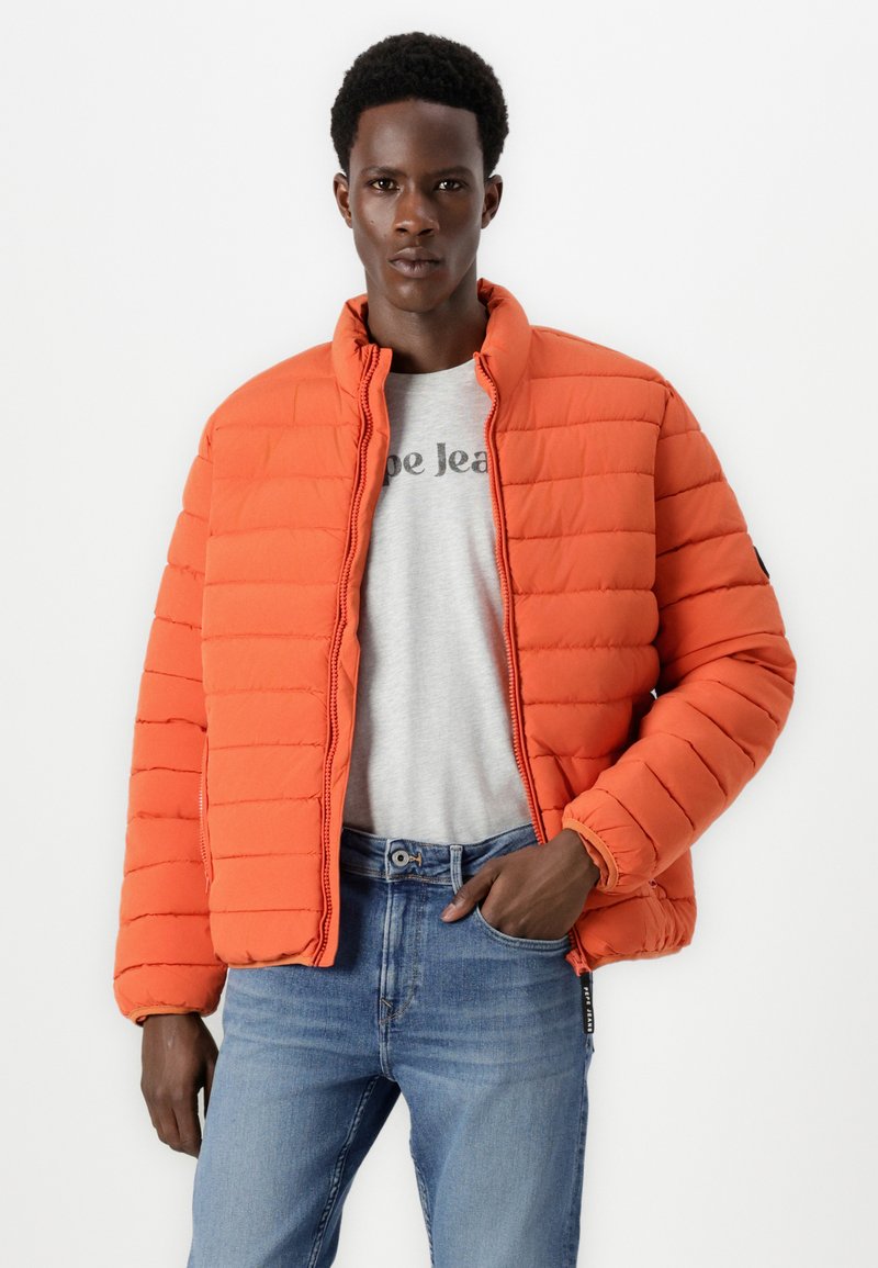 Chaqueta de plumas roja con patrón cuadrado acolchado, con cierre delantero y cuello alto, llevada sobre una camiseta gris y jeans de mezclilla azules.