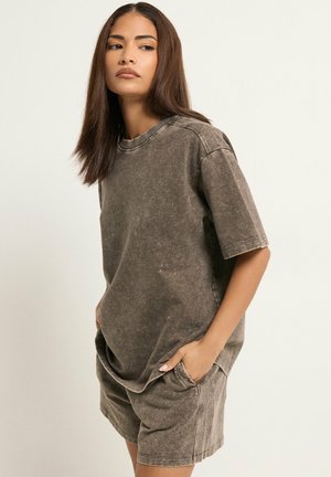 Donna con capelli castani di media lunghezza che indossa una maglietta marrone slavata oversize e pantaloncini abbinati, mani in tasca, guarda leggermente di lato.