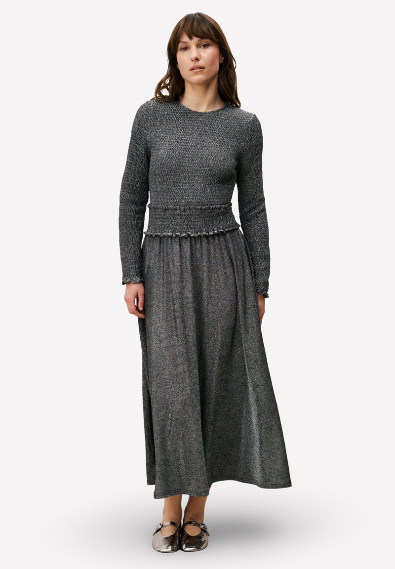 Robe maxi en tissu gris texturé avec un corsage ajusté et une jupe évasée, comprenant un détail volanté à la taille et des manches longues, associée à des chaussures argentées.