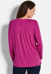 Langärmliges Oberteil aus leuchtend pinkem Stoff, mit lockerer Passform, Rückenschlitzdetails und rundem Halsausschnitt. Kombiniert mit dunkelblauen Hosen.