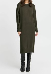 Abito tricot verde oliva con maniche lunghe, polsini a coste e silhouette dritta, abbinato a stivali neri aderenti fino al ginocchio.