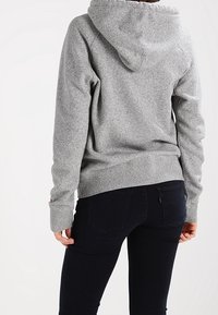 Felpa grigia in pile con cappuccio, polsini e orlo a costine. La parte posteriore mostra maniche a raglan e una texture liscia, abbinata a jeans scuri.