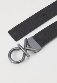 Schwarzer Ledergürtel mit strukturierter Oberfläche, ausgestattet mit einer metallischen Schnalle, die als kreisförmiges "CK"-Logo gestaltet ist. Akzentbeschläge in mattem Grauton.