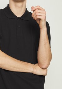 Polo noir avec col classique, patte de boutonnage à deux boutons et manches courtes. Confectionné en tissu lisse avec une texture subtile.