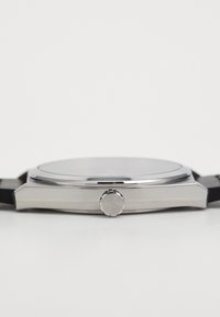 Reloj de tono plateado con un dial redondo, correa de goma negra y una corona texturizada etiquetada como 'HUGO'. Perfil delgado y diseño minimalista.