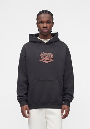 Jeune homme aux cheveux tressés portant un sweat à capuche noir avec un texte graphique orange et un pantalon blanc, debout devant un fond uni.