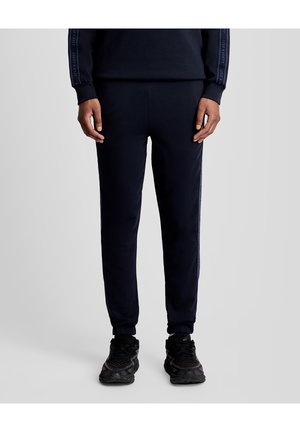 Person trägt schwarze Jogginghosen mit seitlichen Streifen und schwarze Turnschuhe, steht vor einem hellgrauen Hintergrund.