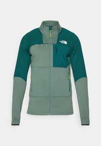 Green outdoor jacket featuring a textured fabric, high collar, front zipper, and contrasting color panels with a chest pocket and logo.